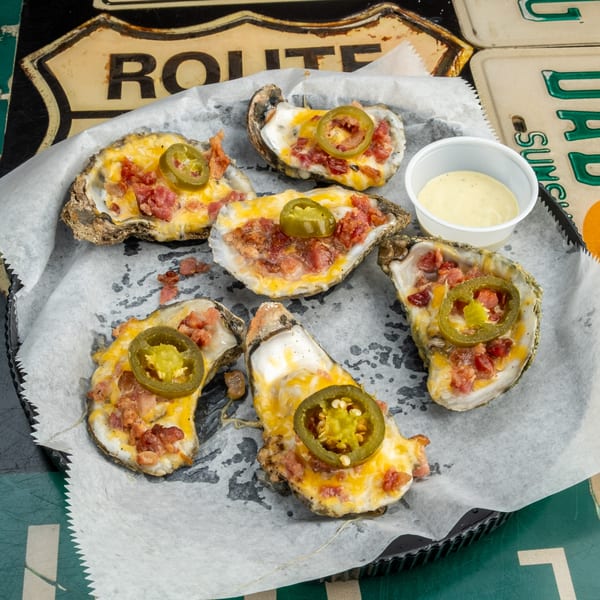 Six baked oysters topped with melted cheese, bacon, and jalapeños, served with a side of sauce.