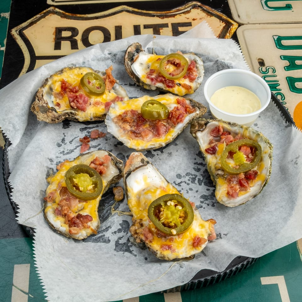 Six baked oysters topped with melted cheese, bacon, and jalapeños, served with a side of sauce.