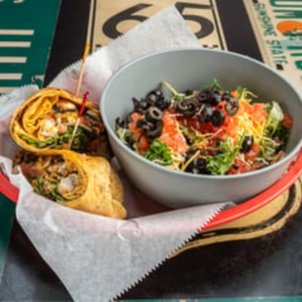 Two wraps in a basket with a side salad topped with olives, tomatoes, and cheese.